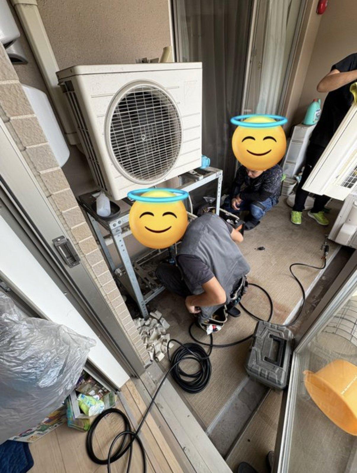 マンションのベランダで、架台に載せた室外機の配管をつなぐ職人と、横でしゃがんで手元を見る人