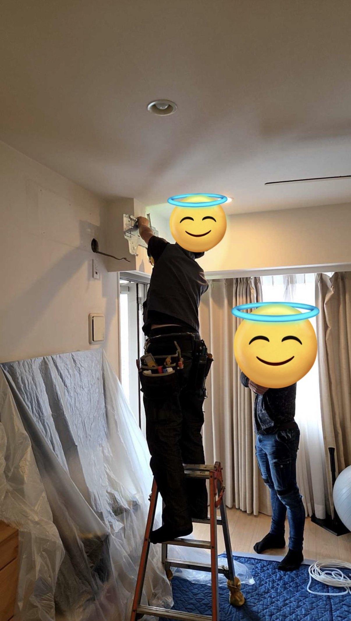 養生シートで家具を覆った部屋で、脚立に乗って室内機の据付板を壁に取り付ける職人と、下で見上げる人